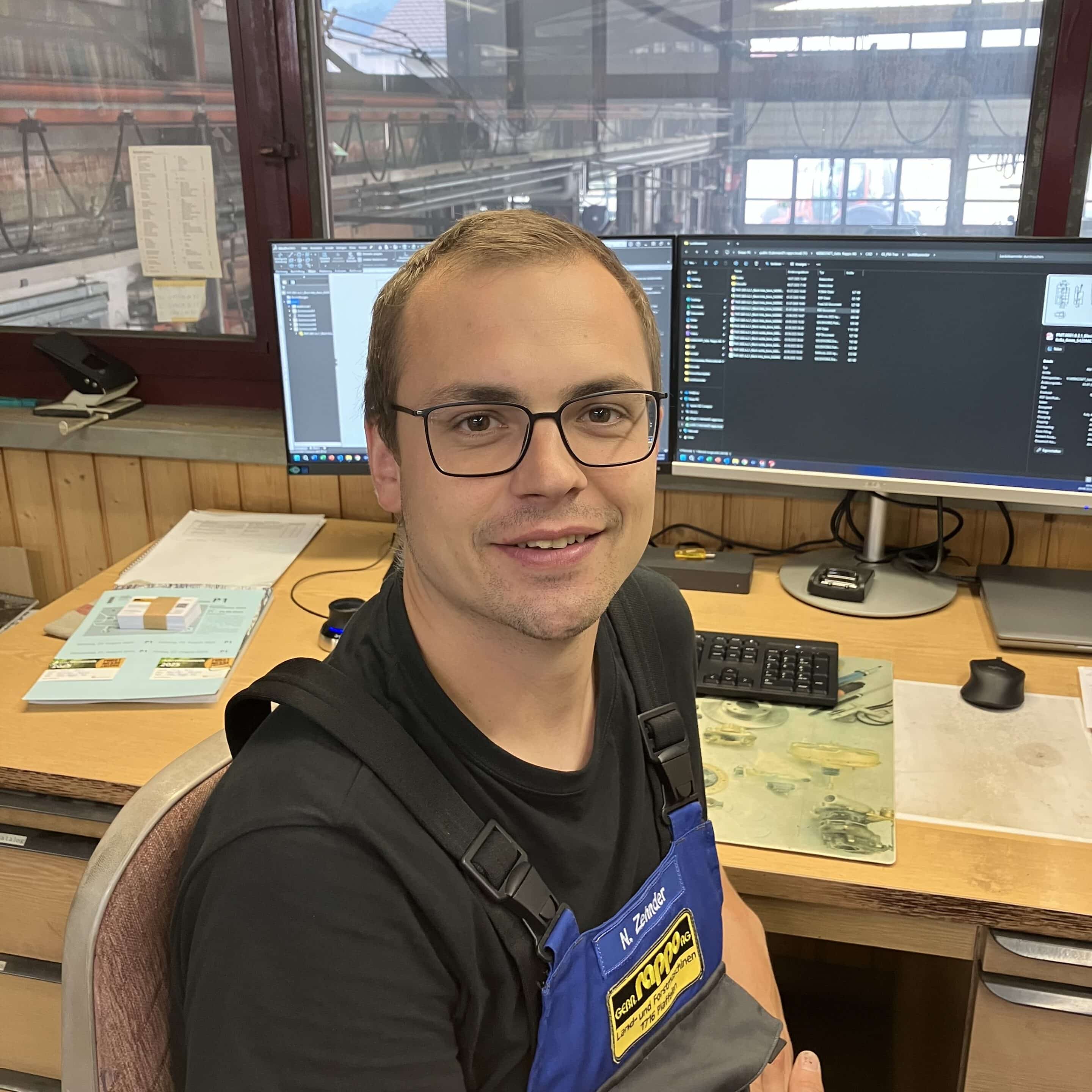 IMG_E5238 Mann im Büro mit Brille und blauer Arbeitskleidung, lächelnd am Schreibtisch mit Computerbildschirmen sitzend.