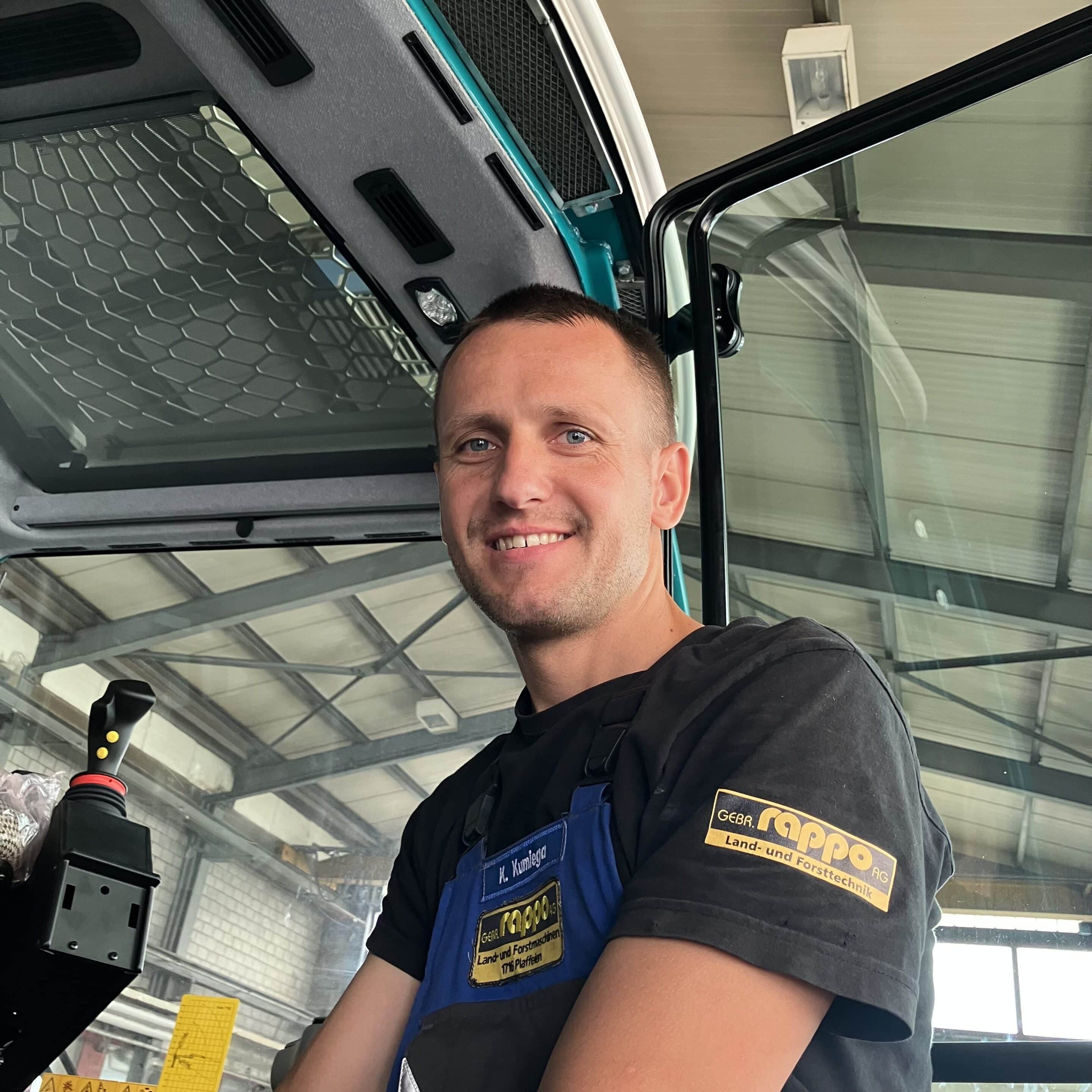 IMG_E5239 Mann in einer Werkstatt lächelt aus der Kabine eines Fahrzeugs mit Firmenaufdruck, porträtiert bei der Arbeit.