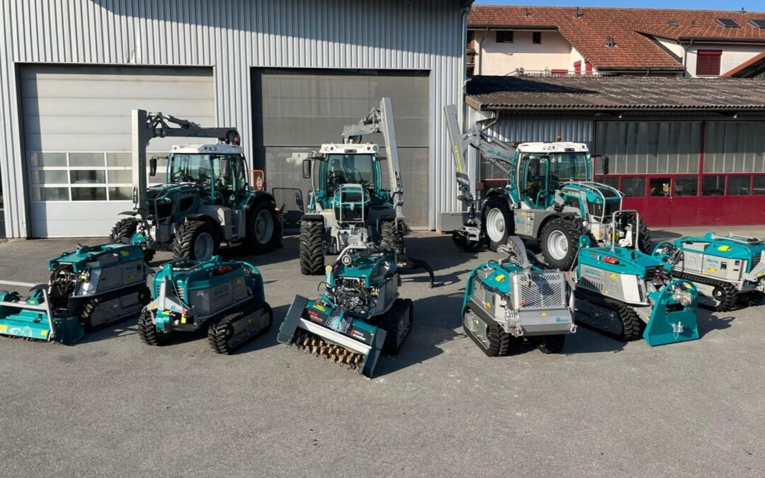 Verschiedene Landmaschinen und Roboter vor einem Firmensitz, sonniger Tag, moderne Landwirtschaftstechnologie.
