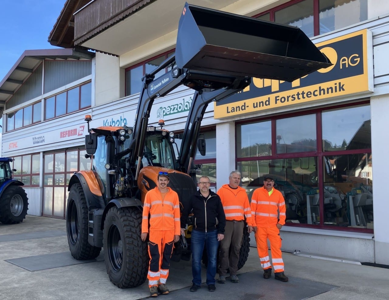 1760338680post_image16 Männer in Arbeitskleidung vor einem großen Traktor bei einem Landtechnik-Geschäft.