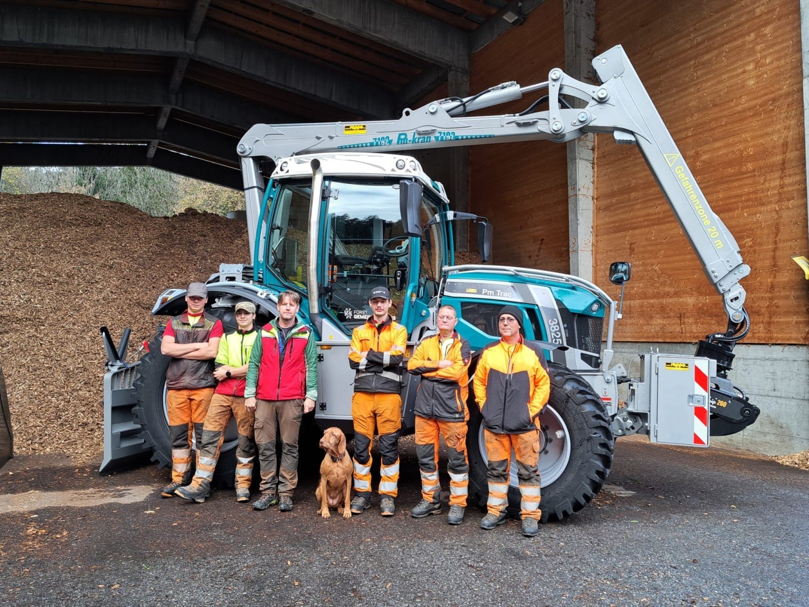 Forstarbeiter-Team und Hund vor einem modernen Traktor mit Kran in Holzbearbeitungsanlage.