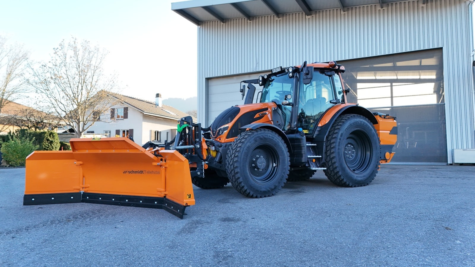 Orange Schneepflugtraktor vor einem Gebäude, bereit für den Winterdienst auf verschneiten Straßen.
