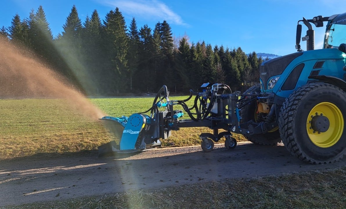 Traktor mit Anbaugerät bearbeitet einen Weg auf einem Feld, Wald im Hintergrund, sonniger Tag.