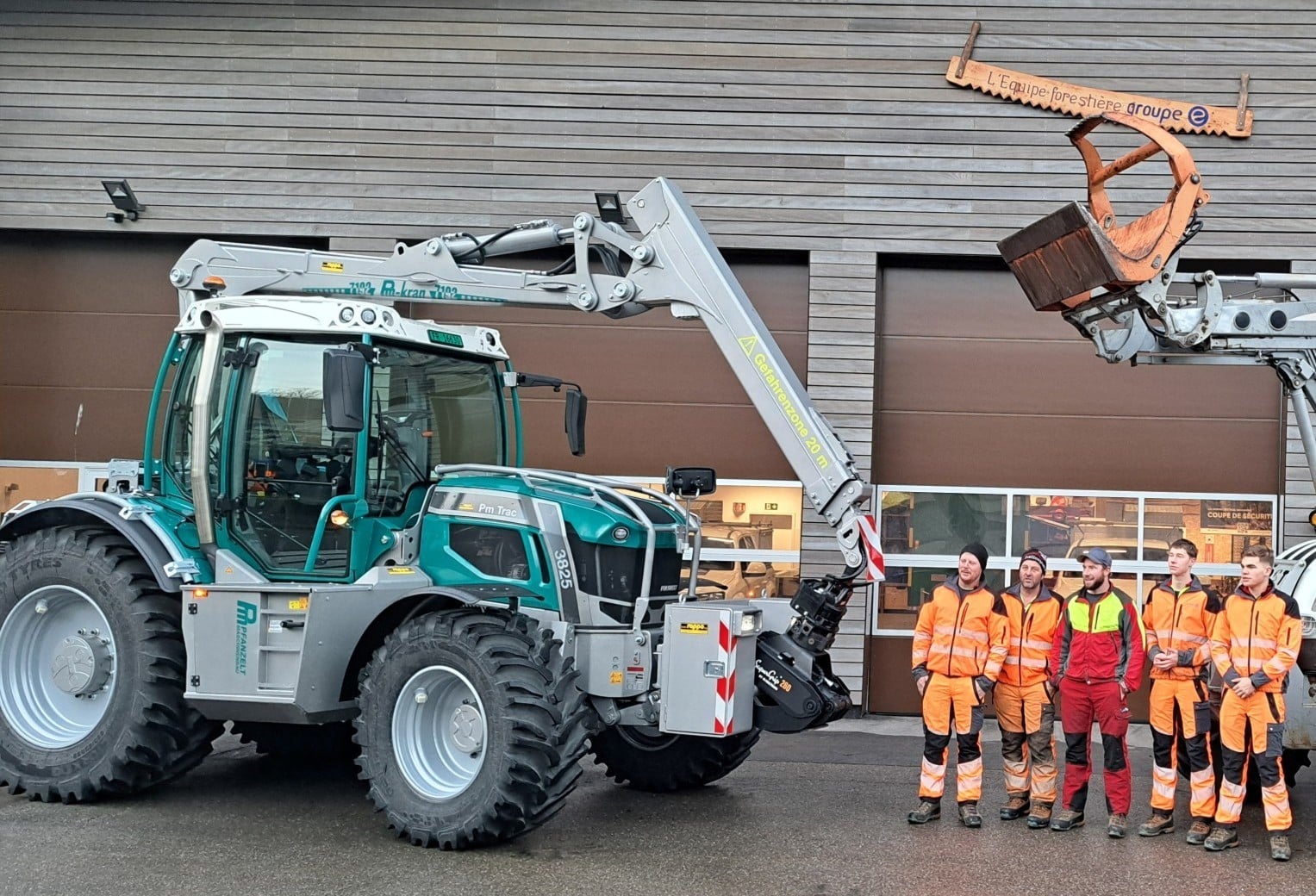 Grüner Traktor mit Kran vor Gebäudefront, fünf Personen in orangefarbener Arbeitskleidung danebenstehend.