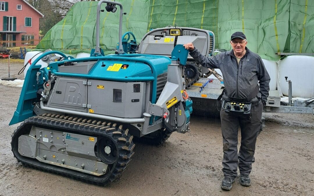 Mann neben einem blauen Raupenfahrzeug auf einem Bauernhof mit grünen Planen im Hintergrund.