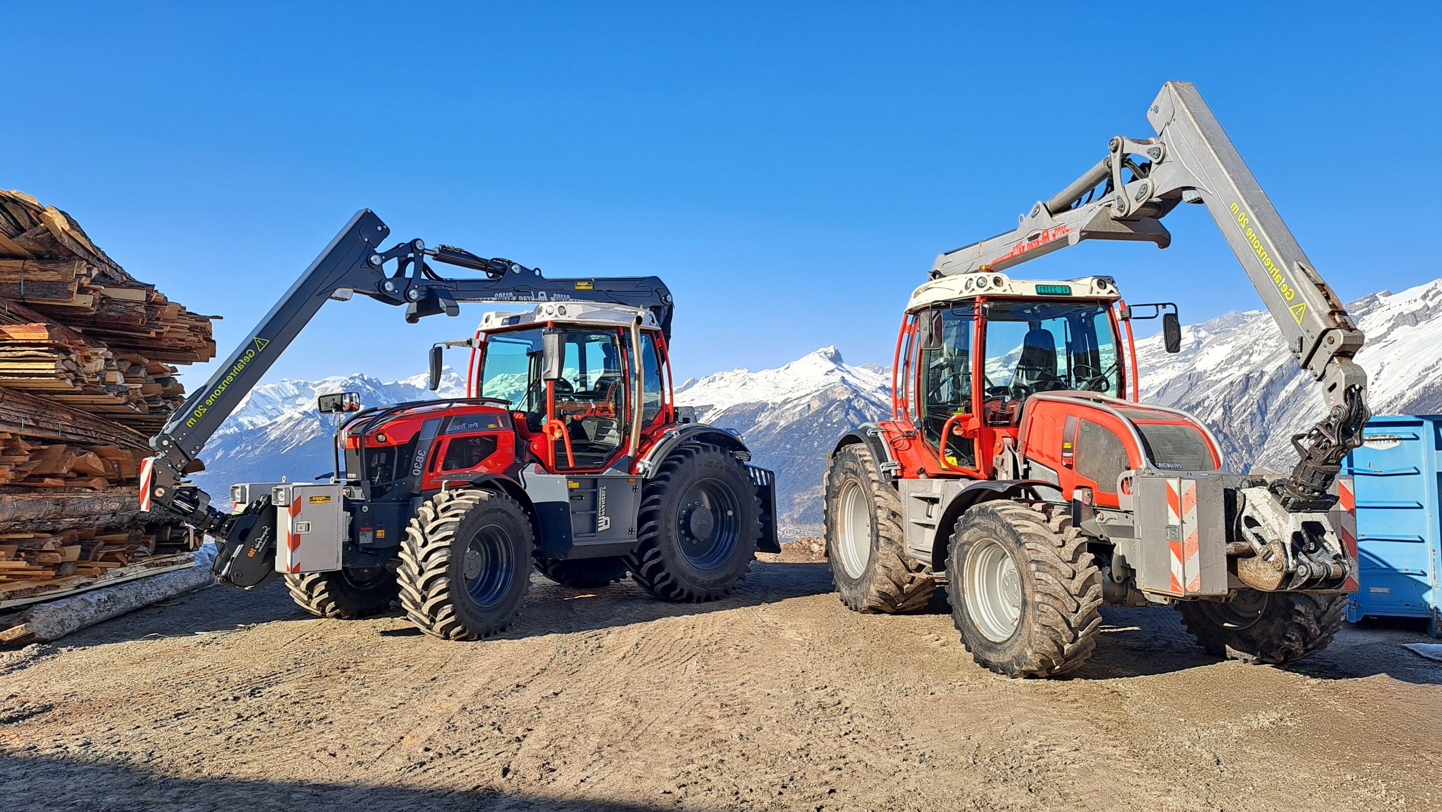 Zwei rote Forsttraktoren mit Greifarmen vor schneebedeckten Bergen stapeln Holzstämme.